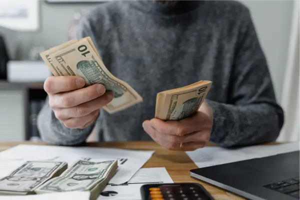 Pessoa contando dinheiro em notas sobre a mesa, com laptop, calculadora e papéis de controle financeiro.