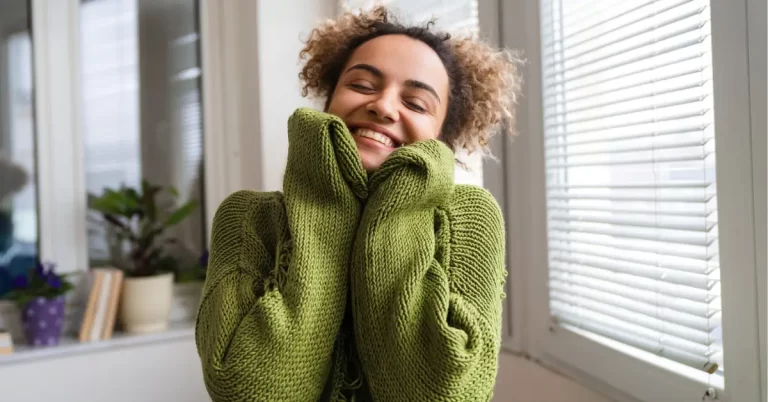 Mulher sorrindo enquanto abraça a si mesma com um suéter verde, transmitindo sensação de amor próprio e bem-estar emocional.