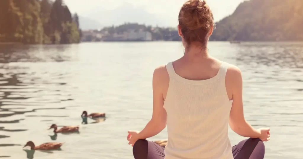 Mulher meditando à beira de um lago ao pôr do sol, simbolizando reflexão interior e perguntas de autoconhecimento.