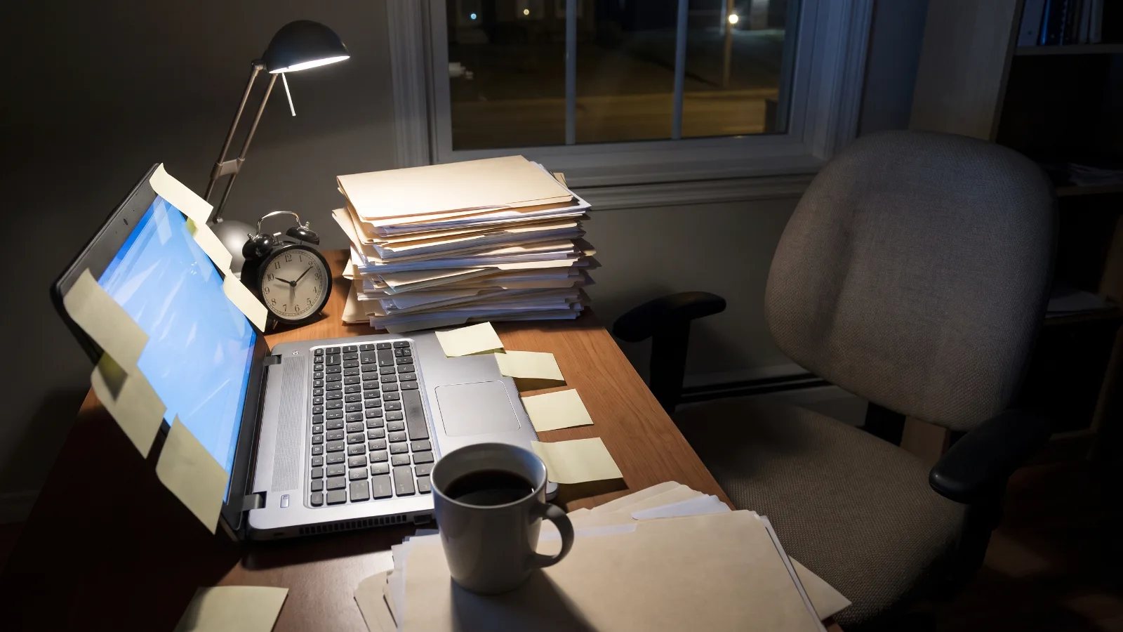 Burnout: o que é na rotina, mesa de trabalho à noite com laptop aceso, pilha de papéis, xícara de café frio e relógio